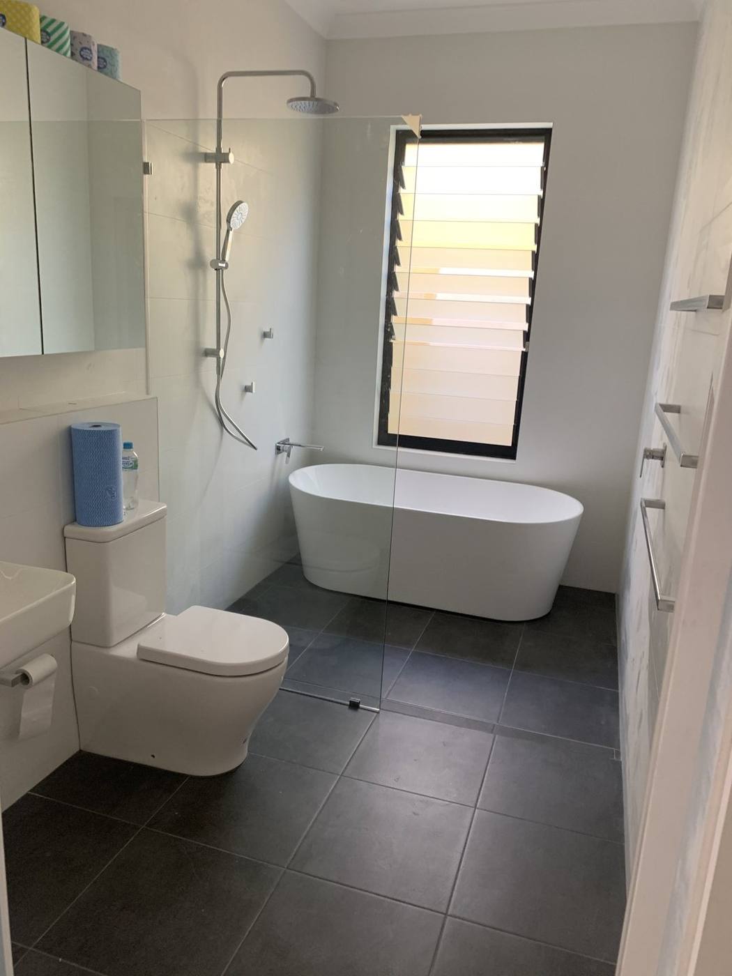 Finished modern bathroom wide view with glass shower screen, freestanding bath, louvre window and dark floor tiles, Dulwich Hill by Loel Homes