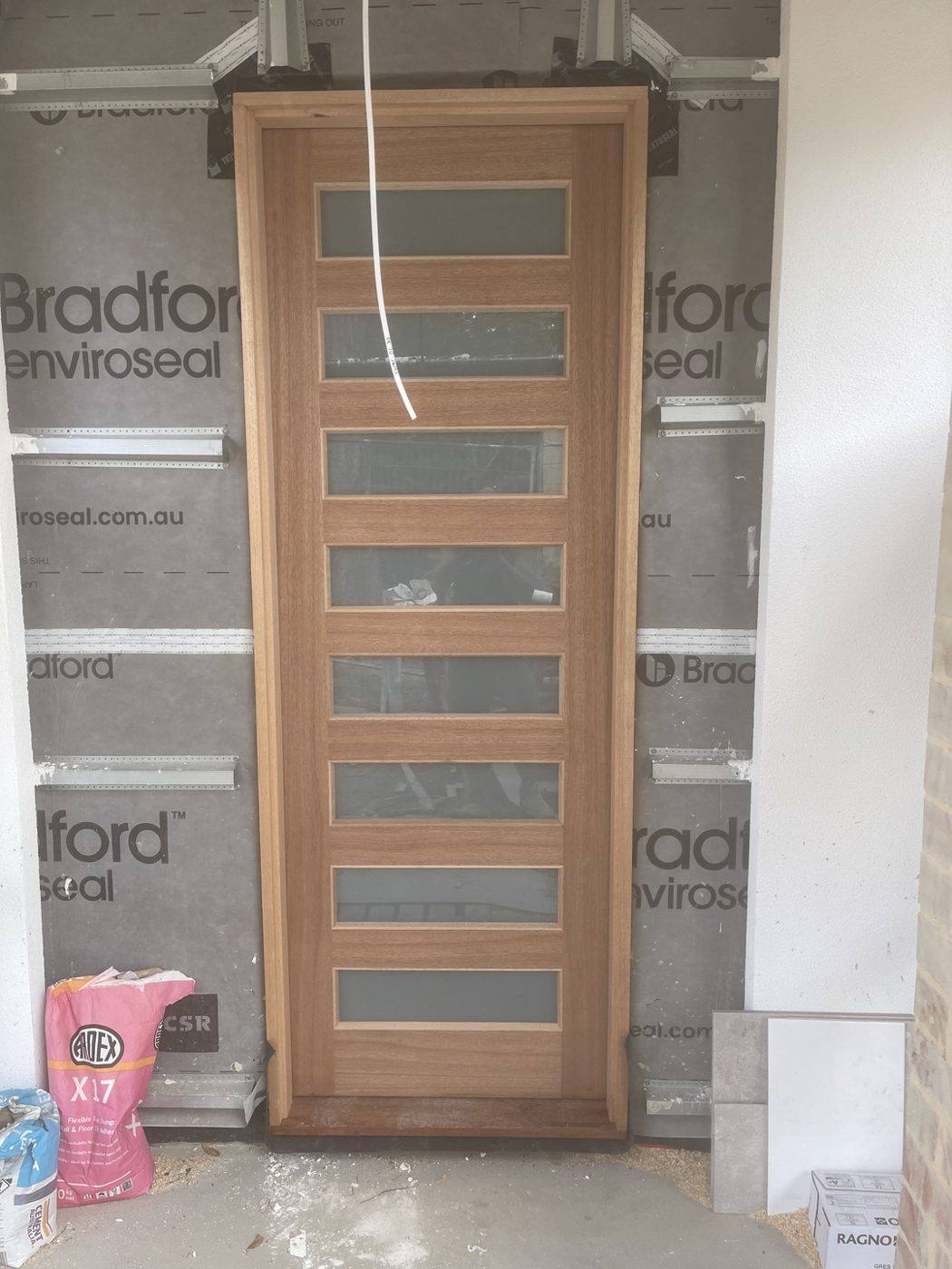 Natural timber front entry door with staggered frosted-glass slots, front view during install by Loel Homes