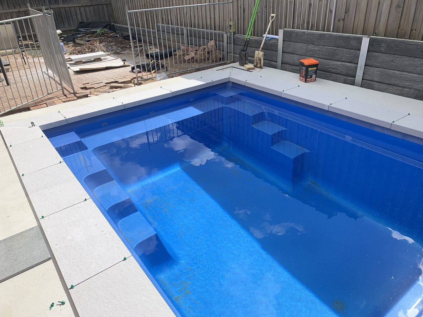 Pool shell filled with stone coping being laid, temporary safety fence still up, by Loel Homes