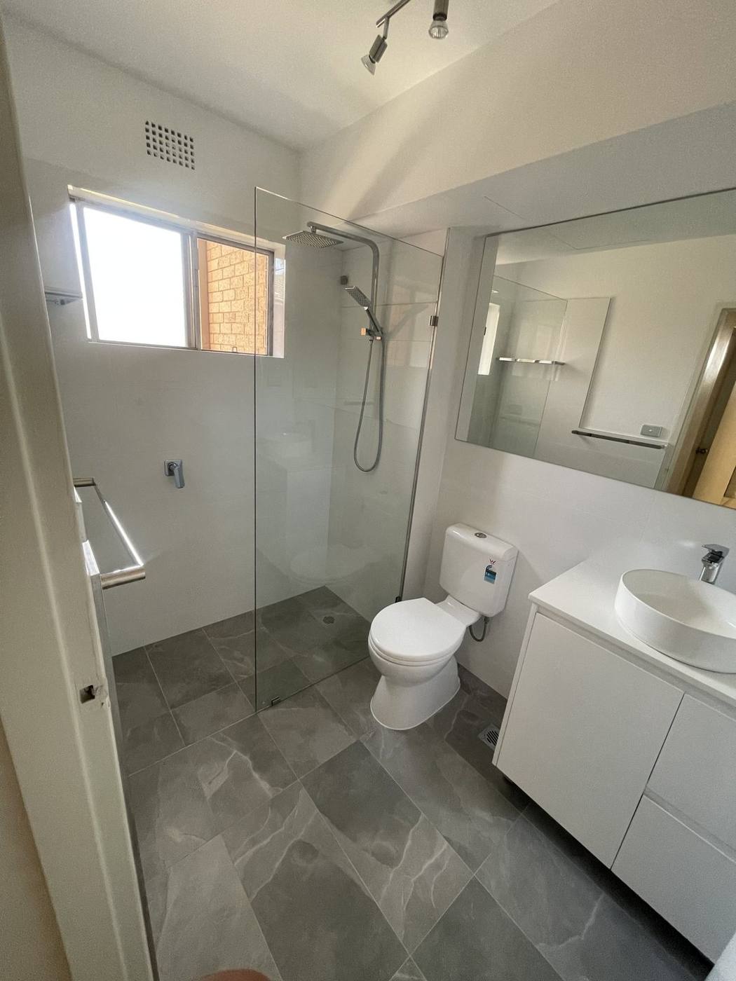 Finished modern bathroom wide view with frameless glass shower, toilet, white vanity, large mirror and grey floor tiles, Top Ryde by Loel Homes